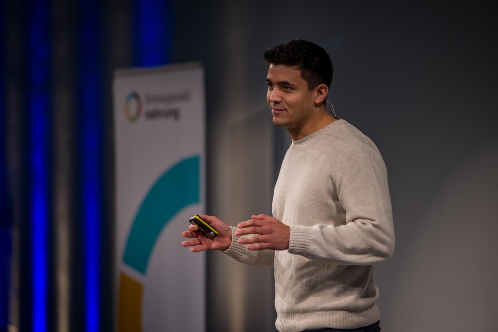 Eine Person in einem hellen Pullover spricht auf der Bühne, hält ein Gerät mit einem Mikrofon-Headset. Im Hintergrund ist ein verschwommenes Banner mit bunten Grafiken zu sehen.