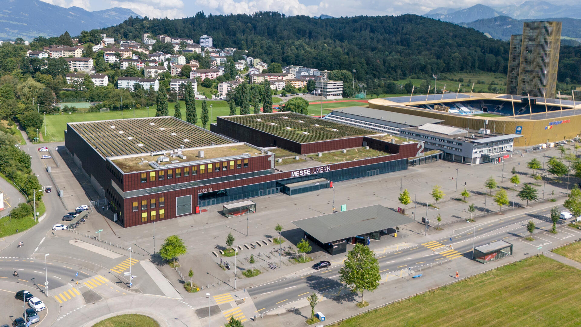 Luftaufnahme des Messezentrums Luzern mit seinen großen rechteckigen Gebäuden, dem Parkplatz und dem angrenzenden Stadion vor einer Kulisse aus grünen Hügeln und verstreuten Wohnhäusern.