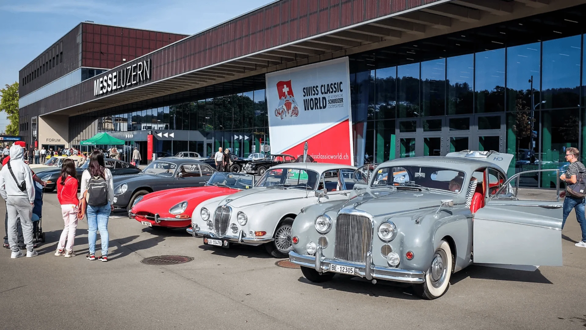 Eine Reihe Oldtimer parkten vor dem Gebäude der Messe Luzern in der Swiss Classic World. Menschen bewunderten die Fahrzeuge und die Glasfenster spiegelten die Szene wider.