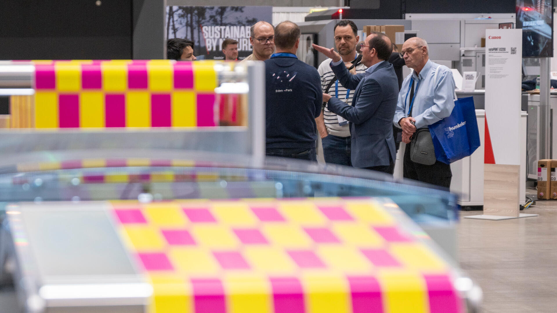Eine Gruppe von Männern in Businesskleidung unterhält sich bei einer Indoor-Veranstaltung. Im Vordergrund sind große, farbenfrohe, rosa und gelb bedruckte Blätter zu sehen. Im Hintergrund sind Ausstellungsstände und Informationstafeln zu sehen.