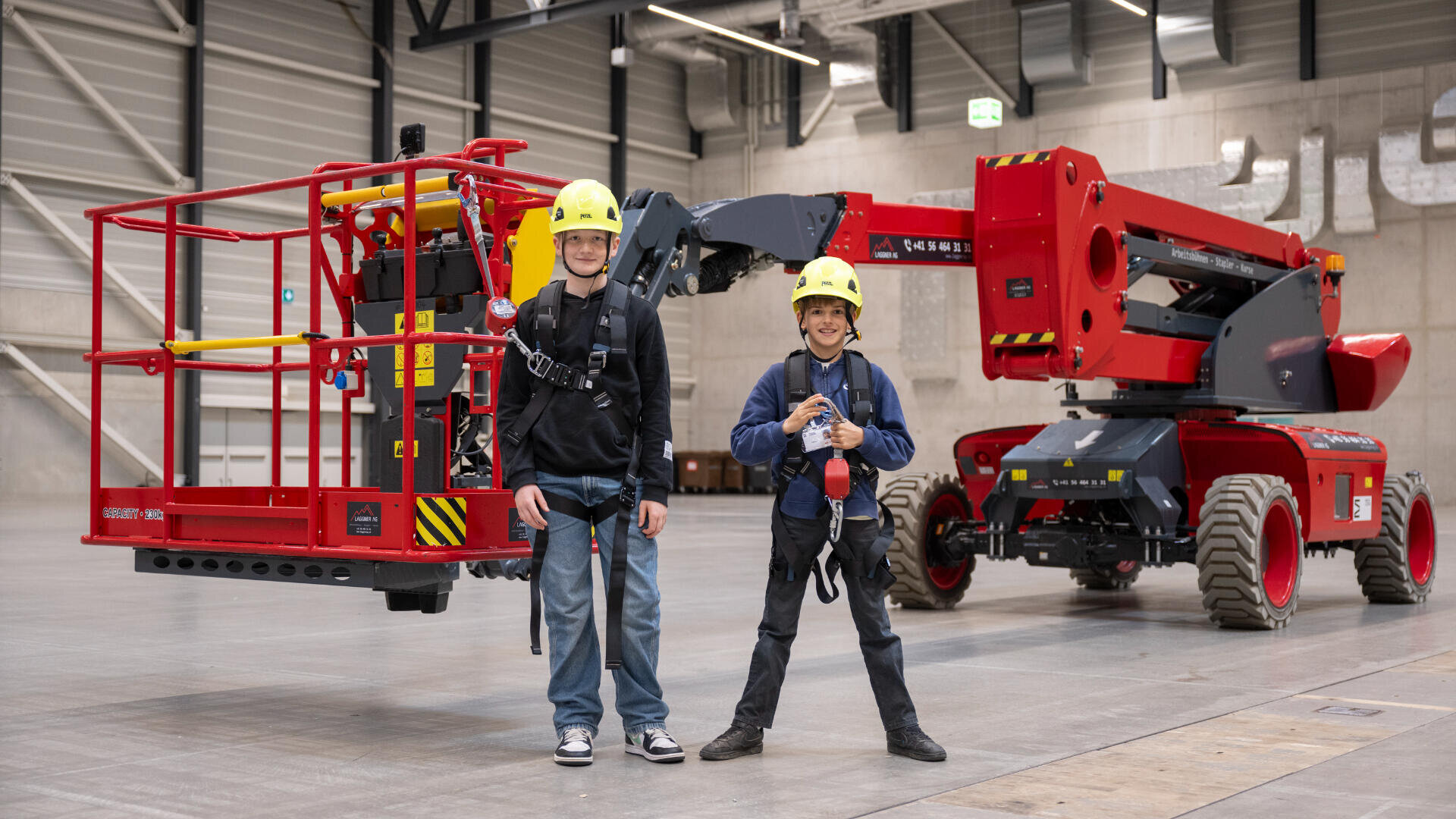 Zwei Kinder mit Schutzhelmen und Gurten stehen lächelnd vor einer großen roten Hebebühne in einem geräumigen Industriegebäude mit Betonwänden und hohen Decken.