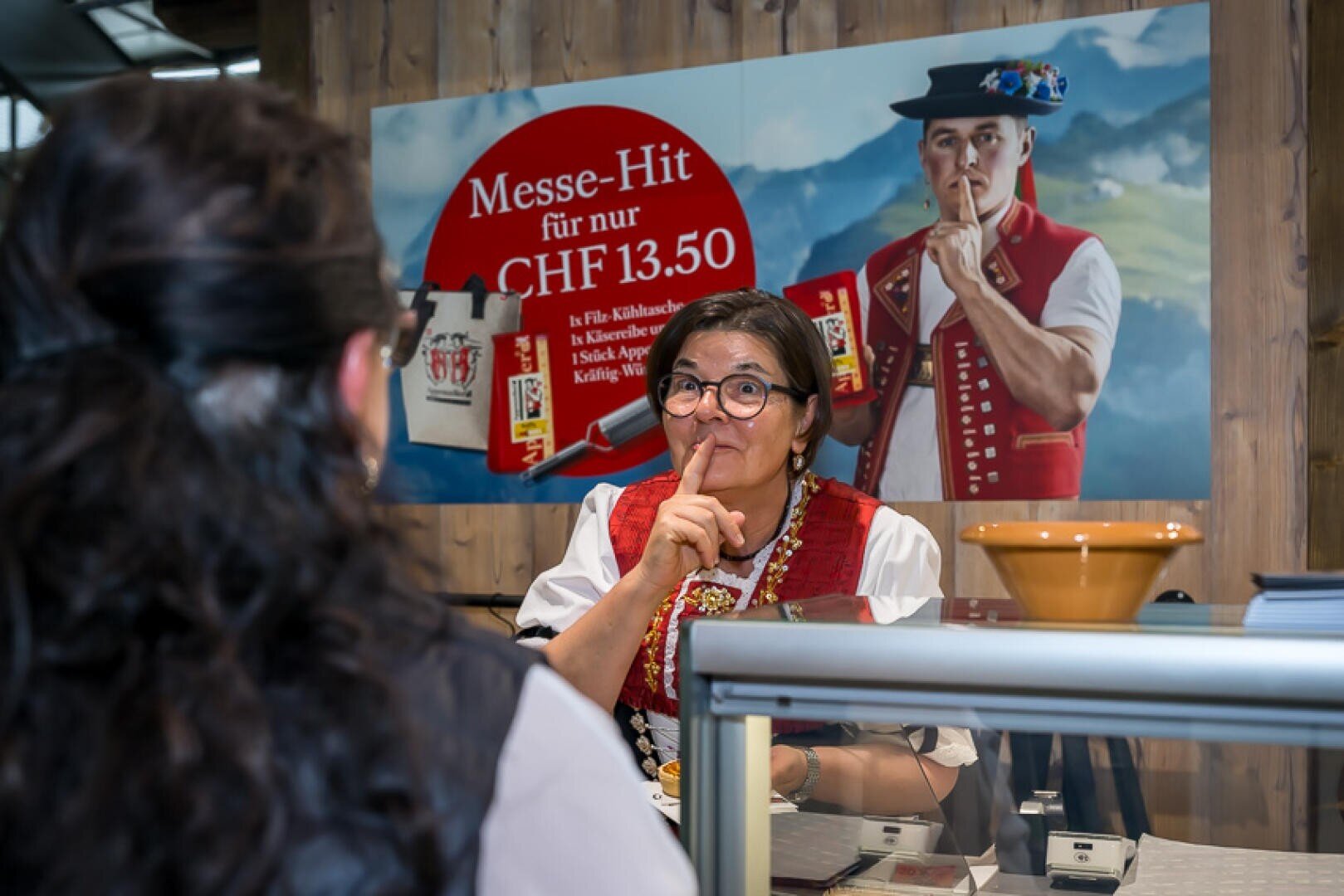 Eine Frau in traditioneller Schweizer Tracht fordert an einem Schalter mit einer Geste Ruhe; dabei ahmt sie einen Mann nach, der auf einem Plakat hinter ihr dieselbe Geste macht, auf dem in deutscher Sprache für ein Sonderangebot geworben wird.