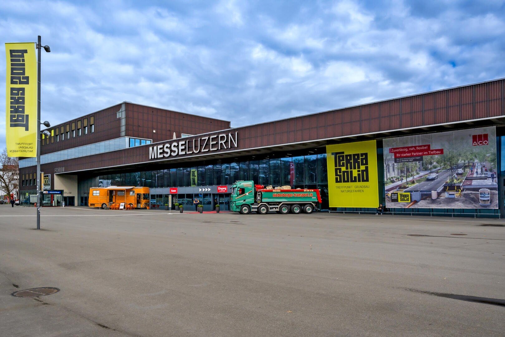 Das Äußere der Messe Luzern, eines Kongresszentrums in Luzern, Schweiz, mit Werbebannern für Veranstaltungen. Lastwagen und ein orangefarbenes Fahrzeug sind vor dem Gebäude geparkt, während der Himmel bewölkt ist.