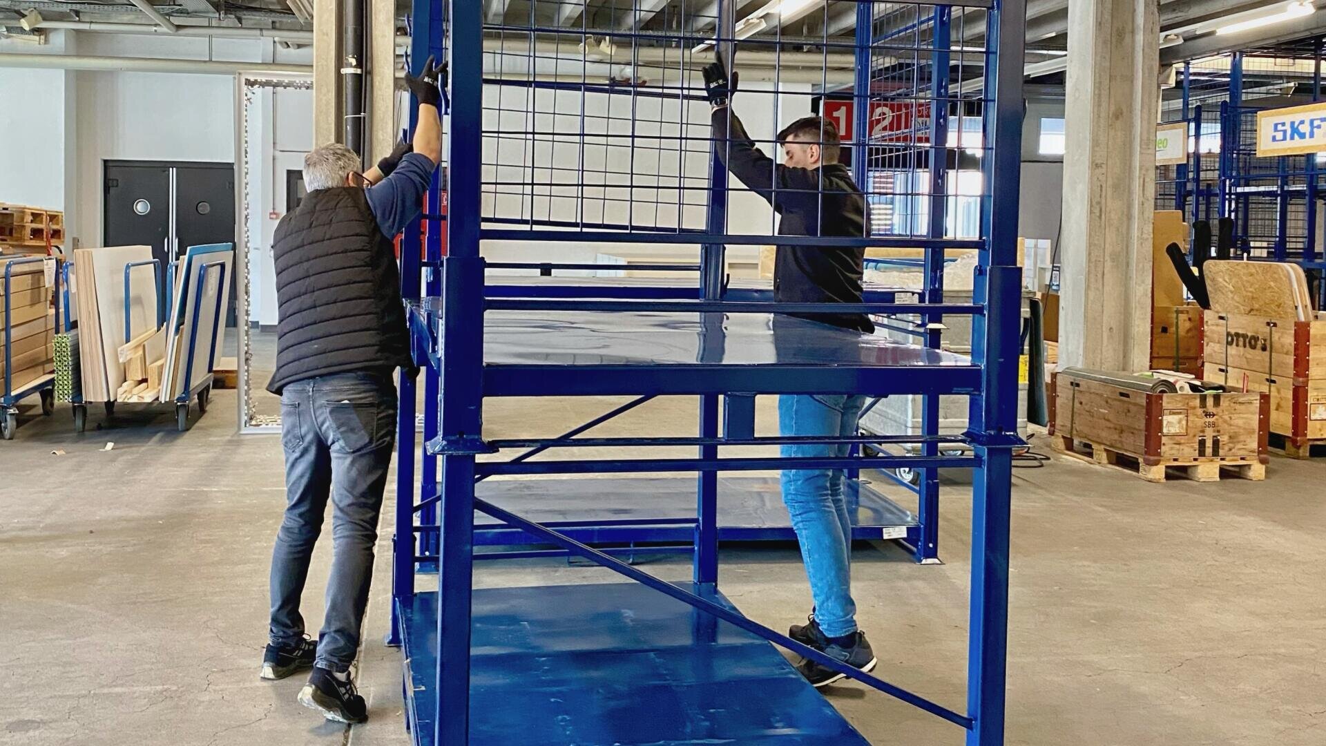 Zwei Personen in Freizeitkleidung und Handschuhen schieben ein großes blaues Metallregal in einem Industrielager, das von verschiedenen Geräten und Holzpaletten umgeben ist.