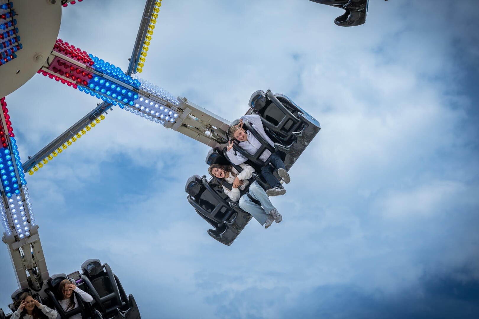 Zwei Menschen lächeln und halten sich an den Händen, während sie auf einer Schaukel im Vergnügungspark hoch in der Luft fahren, mit bunten Lichtern auf der Schaukel und einem bewölkten blauen Himmel im Hintergrund.