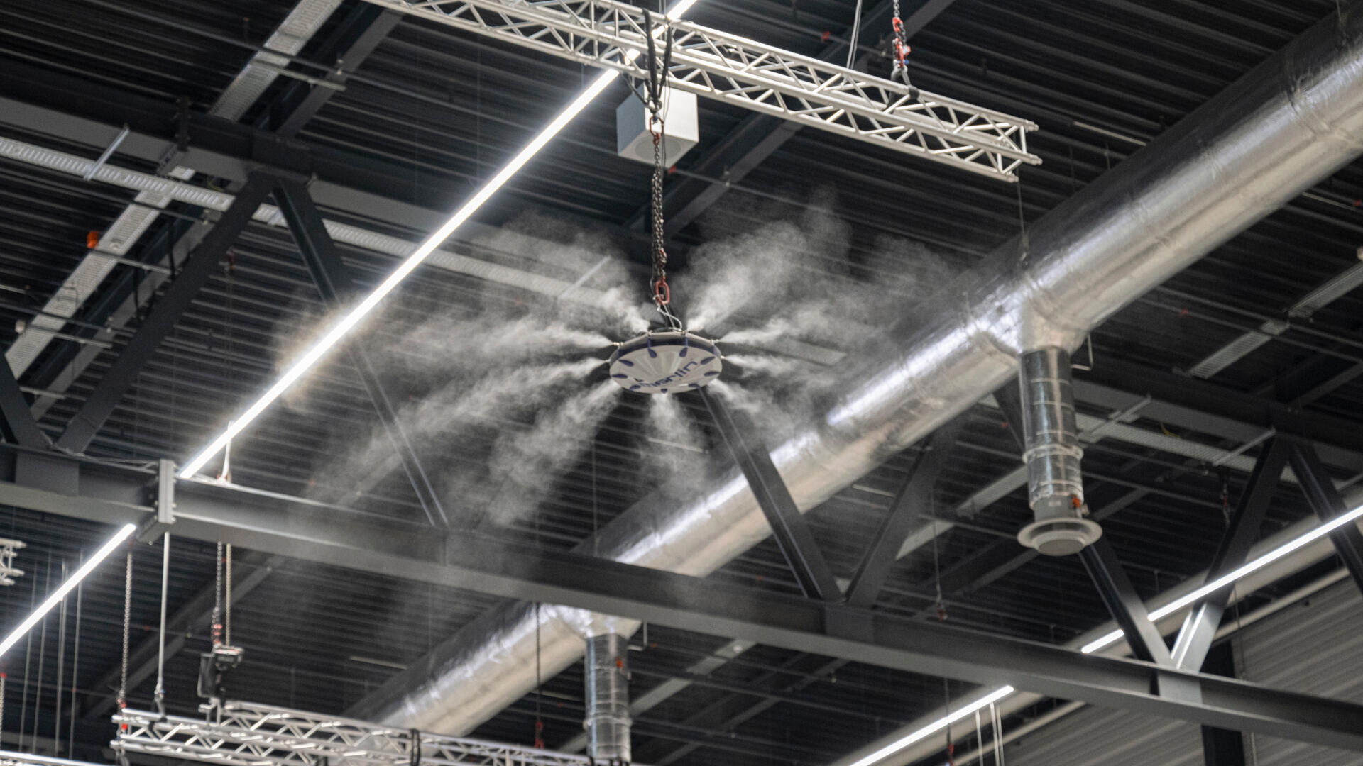 Ein großer Deckenventilator in einem Industriegebäude bläst Nebel in die Luft, umgeben von freiliegenden Kanälen, Rohren und Metallträgern.