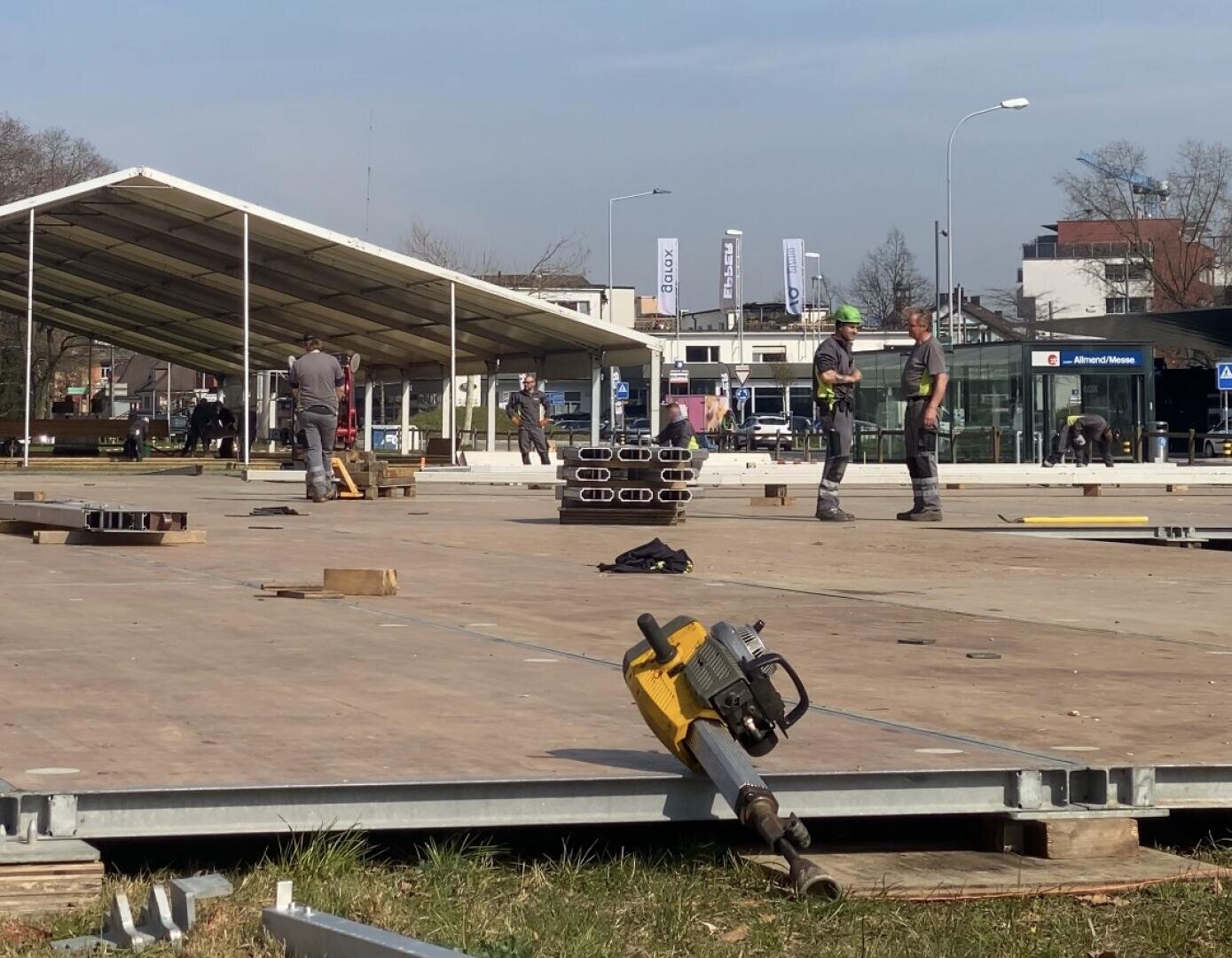 Bauarbeiter montieren eine große Außenstruktur aus Metallträgern und Holzplatten. Im Vordergrund liegt ein Elektrowerkzeug auf dem Boden, und im Hintergrund ist ein überdachter Teil der Konstruktion zu sehen.