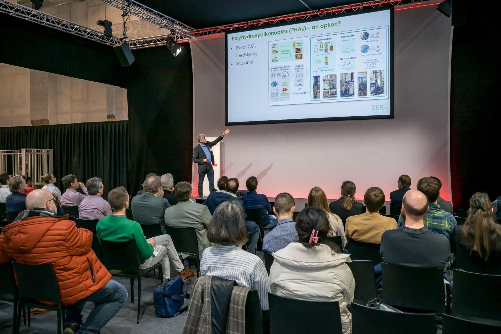 Ein Redner präsentiert einem Publikum in einem Konferenzraum eine wissenschaftliche Diashow. Die Folie behandelt Polyhydroxyalkanoate (PHA) und enthält Diagramme und Text. Das Publikum hört aufmerksam zu.