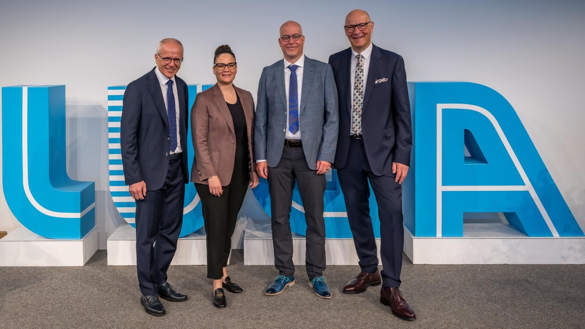 Vier Personen in Geschäftskleidung stehen lächelnd vor großen blauen und weißen Buchstaben, die LUNA auf einer Bühne oder bei einer Veranstaltung buchstabieren.