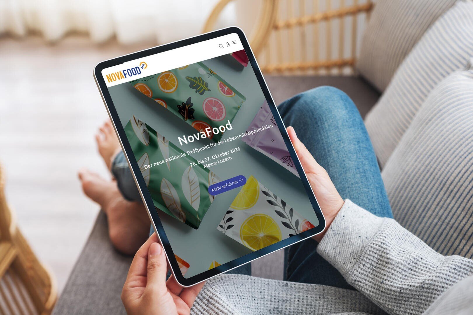 Eine Person hält ein Tablet in der Hand, auf dem die NovaFood-Veranstaltungswebsite mit bunten Fruchtillustrationen und Veranstaltungsdetails angezeigt wird. Die Person sitzt mit gekreuzten Beinen auf einer Couch, die im Hintergrund zu sehen ist.
