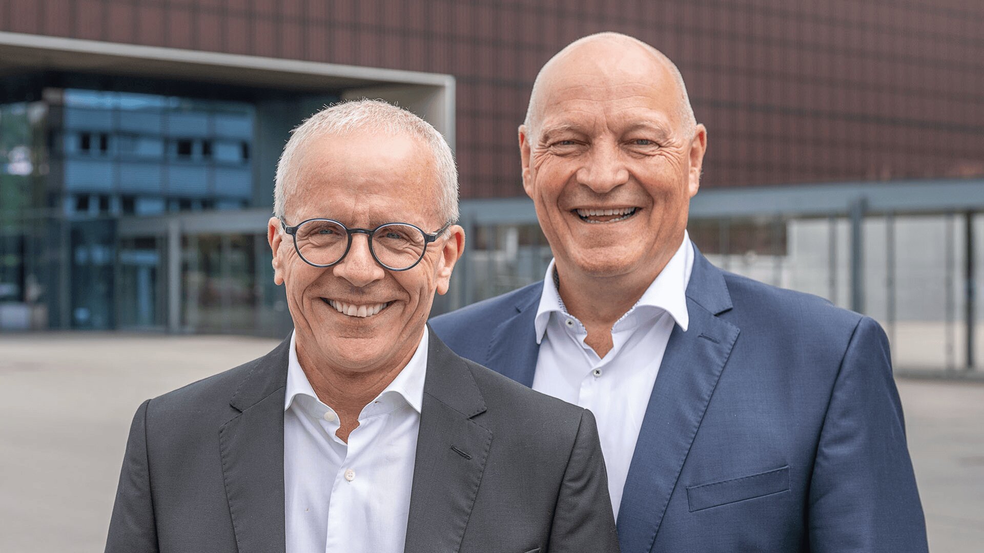 Zwei ältere Männer mit Anzug und Brille stehen draußen vor einem modernen Gebäude und lächeln in die Kamera. Ein Mann steht im Vordergrund, der andere steht etwas hinter ihm.