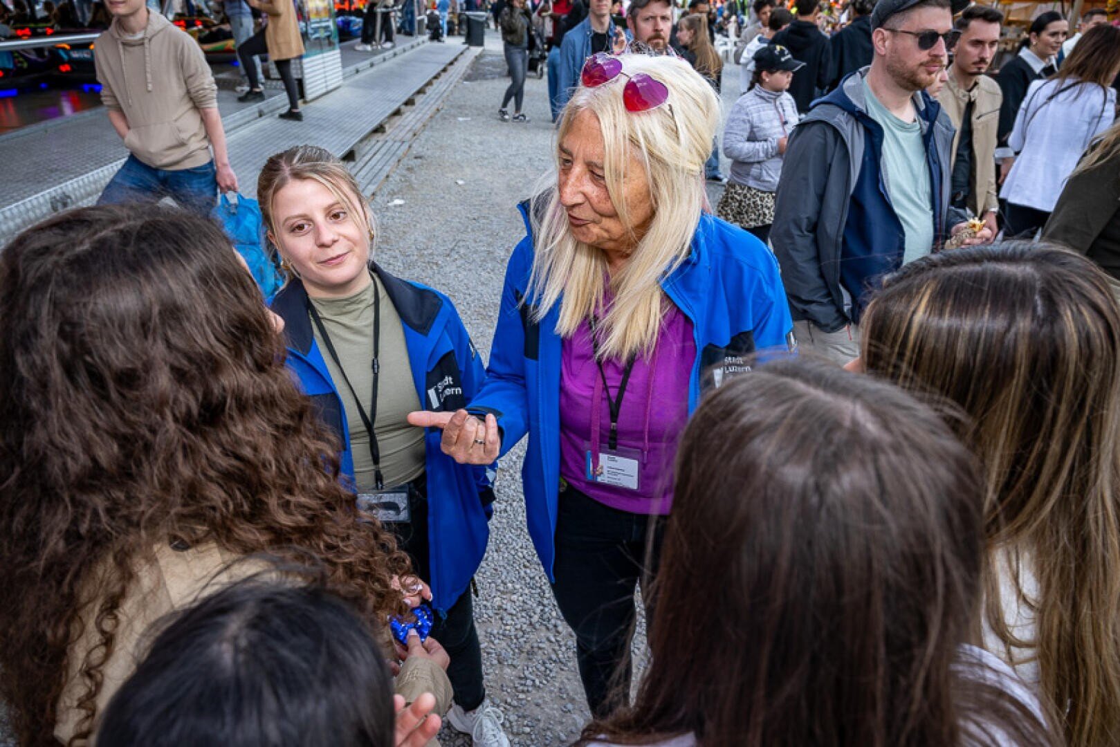 Zwei Frauen in blauen Jacken mit Namensschildern sprechen mit einer kleinen Gruppe von Mädchen bei einer Veranstaltung im Freien, umgeben von Menschen und Aktivitäten im Hintergrund. Eine Frau zeigt auf etwas, während die andere zuhört.