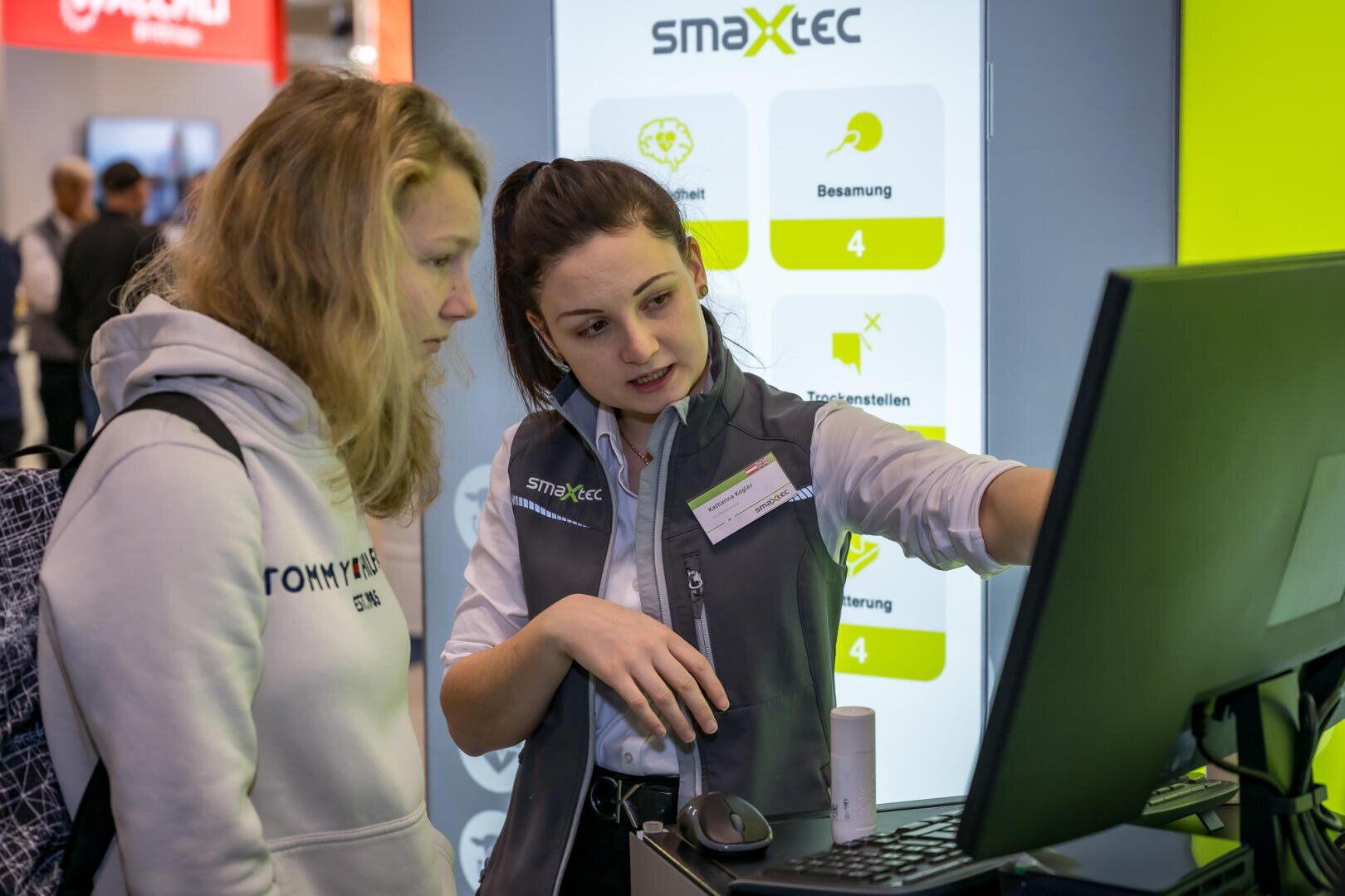 Zwei Frauen stehen vor einem Computermonitor an einem Technikstand; eine Frau zeigt auf den Bildschirm und erklärt der anderen etwas. Im Hintergrund ist ein Display mit dem smaxtec-Logo und Symbolen zu sehen.
