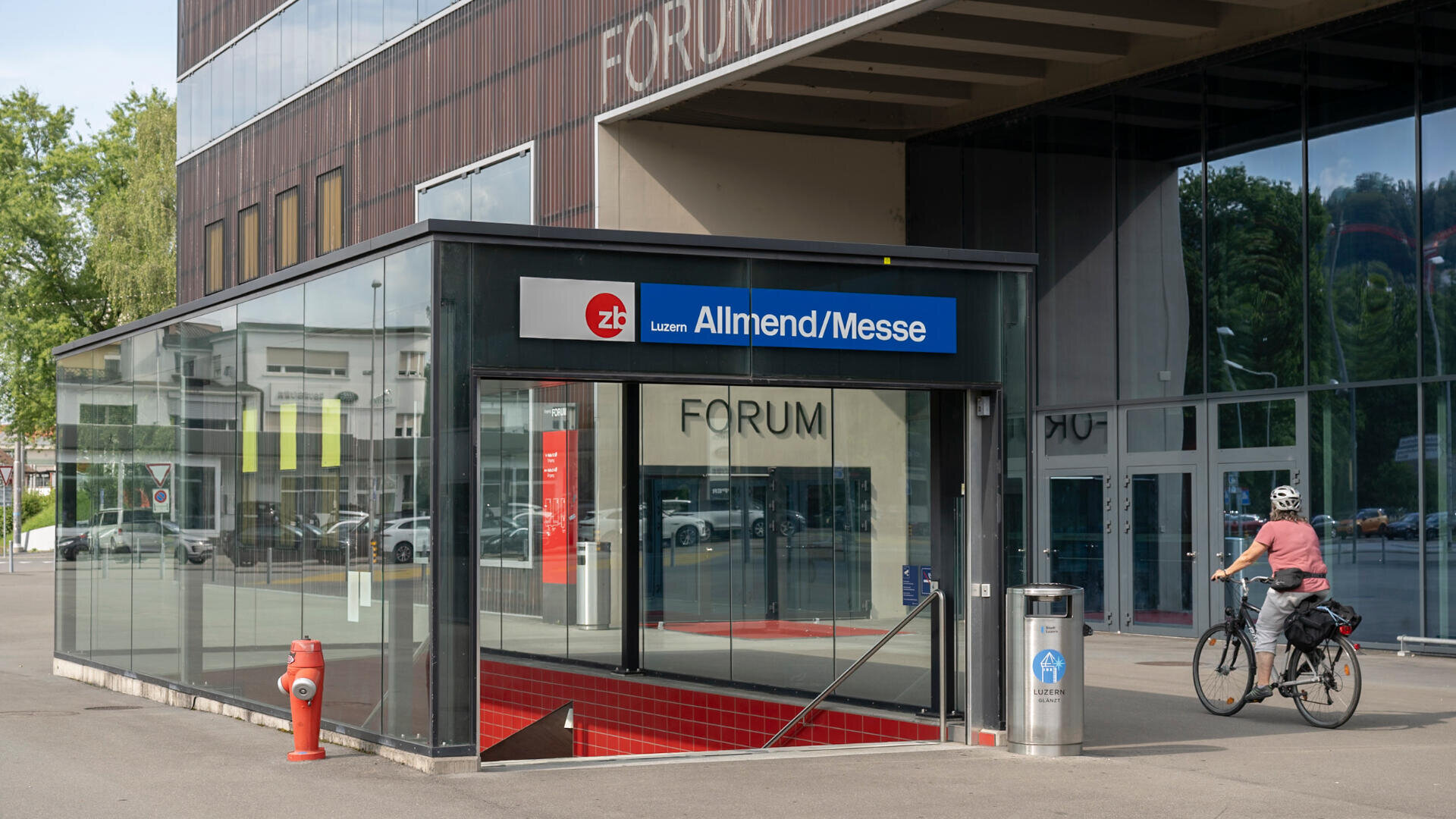 Eingang zum Bahnhof Luzern Allmend/Messe mit Glaskonstruktion, roter Treppe und einem vorbeifahrenden Radfahrer. Im Hintergrund ist das Forum-Gebäude zu sehen.