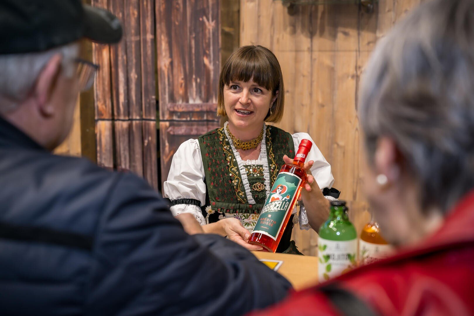 Eine Frau in traditioneller Kleidung überreicht zwei Personen an einem Tisch eine rote Flasche. Auf dem Tisch sind mehrere Flaschen zu sehen, und die Umgebung wirkt rustikal mit Holzwänden im Hintergrund.