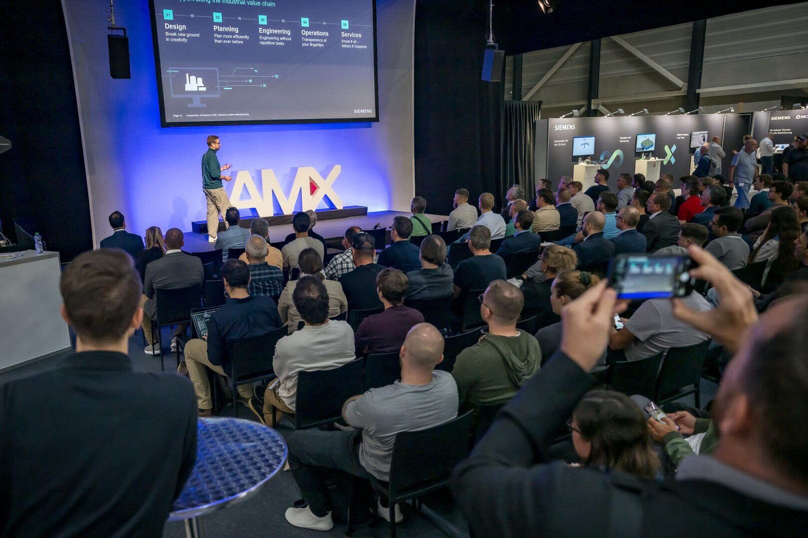 Ein Redner hält einen Vortrag vor einem sitzenden Publikum auf einer Konferenz. Die Zuhörer hören aufmerksam zu, einige machen Fotos, während auf einem großen Bildschirm eine Folie über Design, Planung, Technik, Betrieb und Dienstleistungen gezeigt wird.