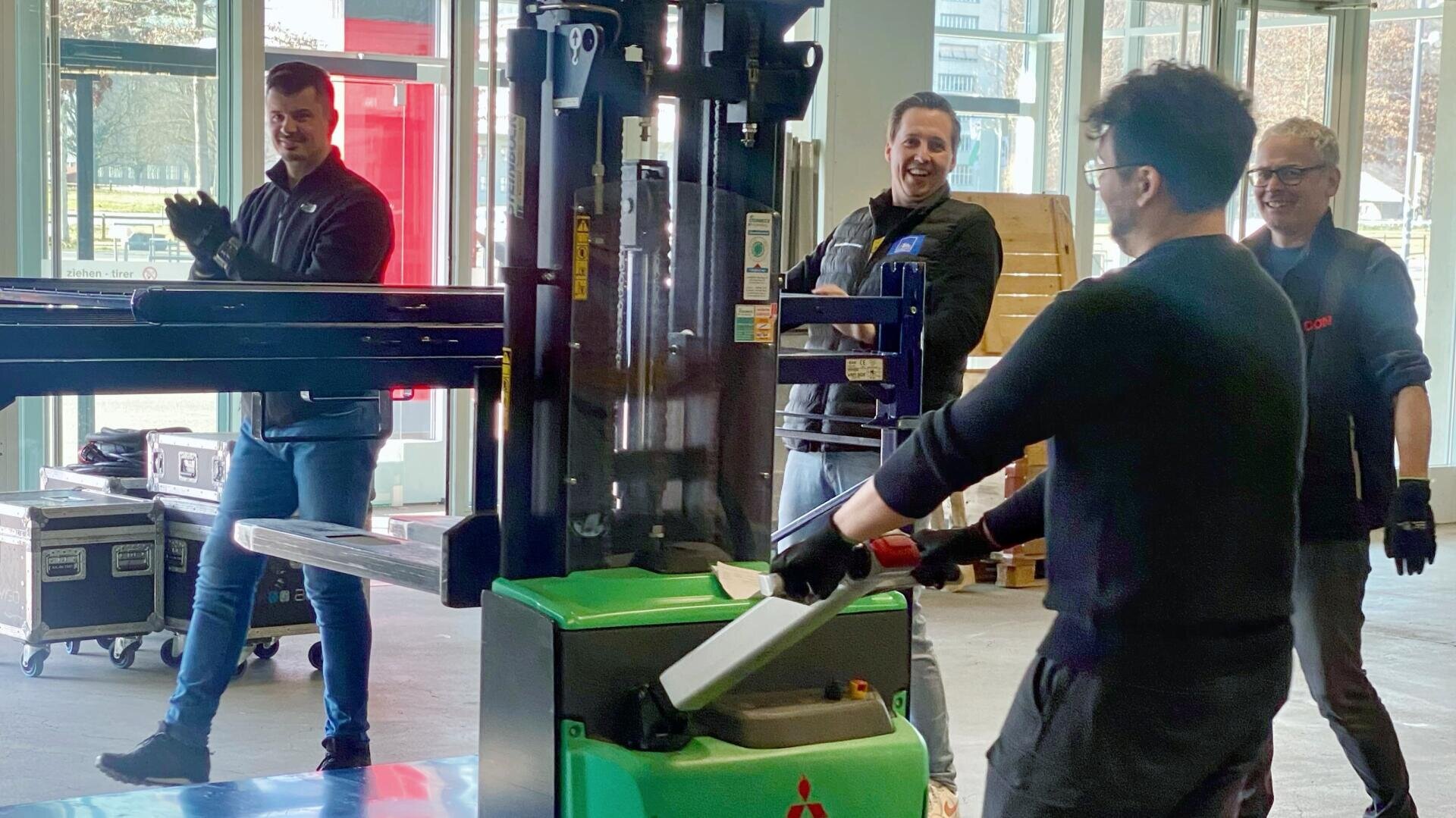 Vier Männer stehen lächelnd und lachend um einen grünen Gabelstapler in einer Halle. Ein Mann bedient den Gabelstapler, während die anderen zusehen und klatschen und so eine lebhafte und fröhliche Atmosphäre schaffen.
