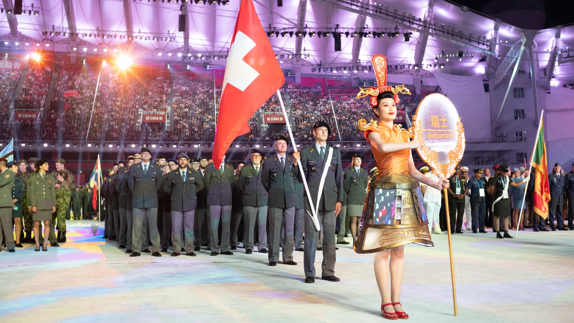 Eine Frau in reich verzierter traditioneller Kleidung führt eine Gruppe uniformierter Schweizer Soldaten an, die eine große Schweizer Flagge halten, während einer Zeremonie in einem hell erleuchteten Stadion voller Zuschauer.