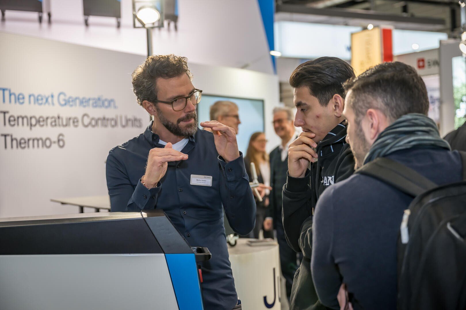 Ein Mann mit Brille erklärt zwei aufmerksamen Männern an einem Technologiestand mit Hilfe von Handgesten etwas. Auf einem Display im Hintergrund ist zu lesen: Die nächste Generation: Temperiergeräte Thermo-6.