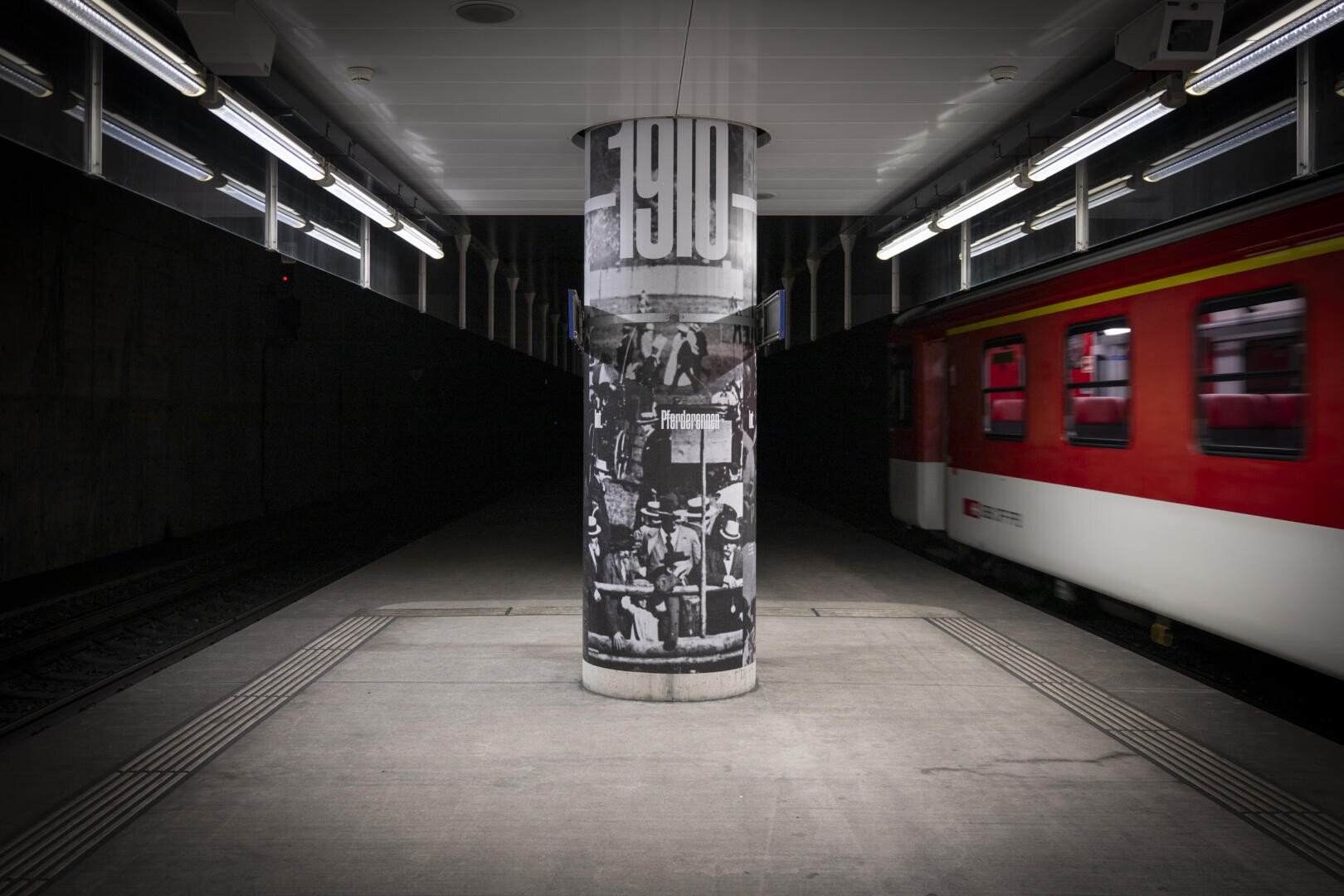 Ein U-Bahnsteig mit einer Säule in der Mitte, auf der Schwarzweißfotos und die Zahl 190 zu sehen sind. Rechts ist teilweise ein roter Zug zu sehen, im Hintergrund ist der Tunnel dunkel.