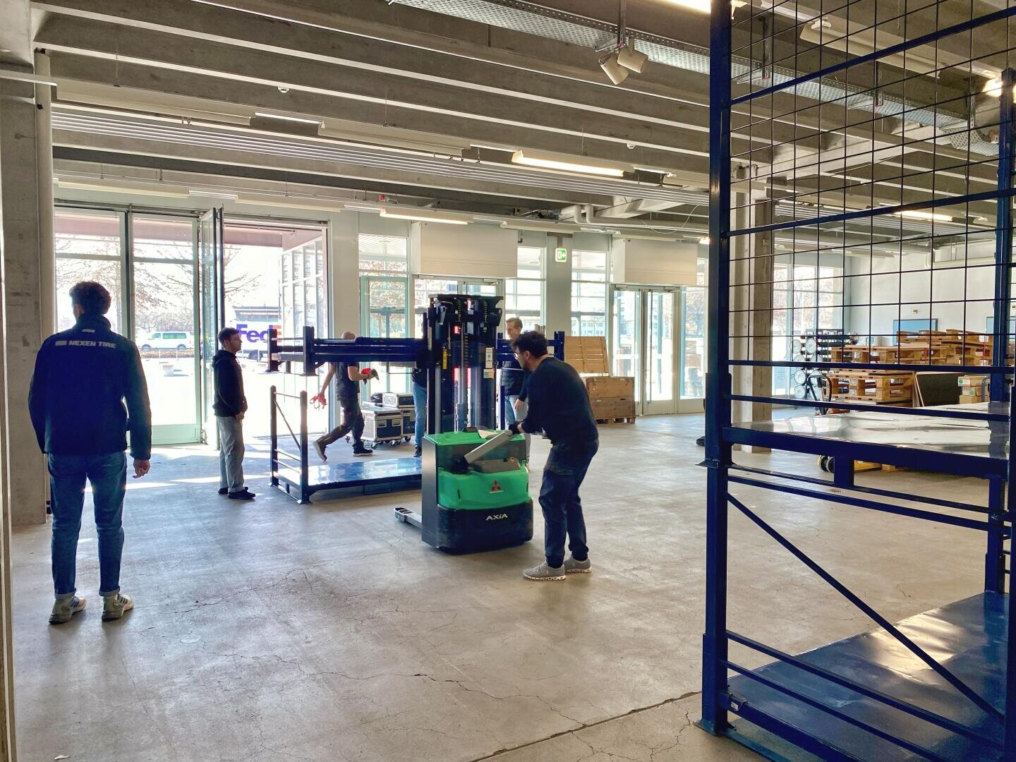 Fünf Personen arbeiten gemeinsam in einem geräumigen Industrielager; einer bedient einen grünen Palettenheber, während die anderen die Arbeit anleiten und überwachen, während draußen durch große offene Türen ein FedEx-LKW zu sehen ist. Leere Regale stehen im Vordergrund.