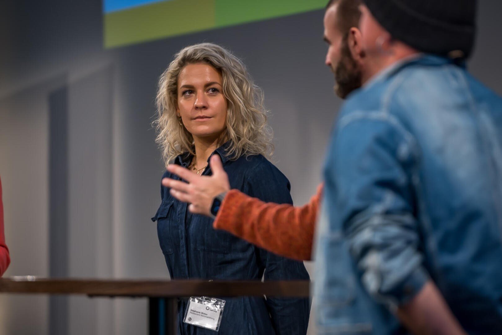Eine Frau mit lockigem blondem Haar hört einem bärtigen Mann in einer Mütze und einer Jeansjacke aufmerksam zu, der während einer Diskussion oder Podiumsveranstaltung beim Sprechen gestikuliert.