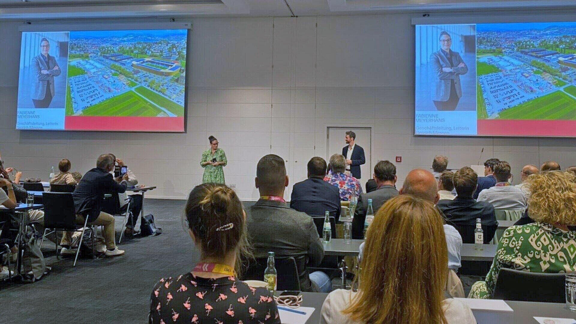 Zwei Redner stehen vorne in einem Konferenzraum und sprechen zu einem Publikum. Eine Folie auf dem Bildschirm zeigt das Foto eines Mannes mit einer Stadtlandschaft im Hintergrund und Text, während die Teilnehmer sitzen und zuhören.