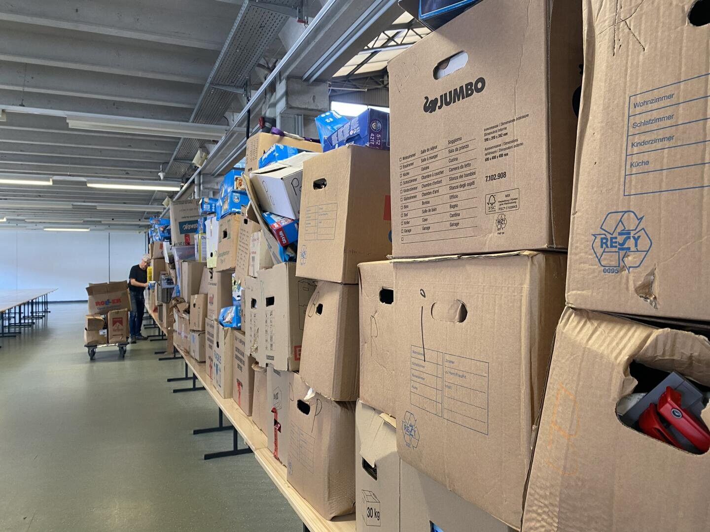In einem großen, hell erleuchteten Raum sind Stapel von Kartons auf Tischen aufgereiht. Im Hintergrund ist eine Person zu sehen, die Kartons sortiert. Die Szene deutet auf einen Umzug, eine Lagerung oder ein Lager hin.