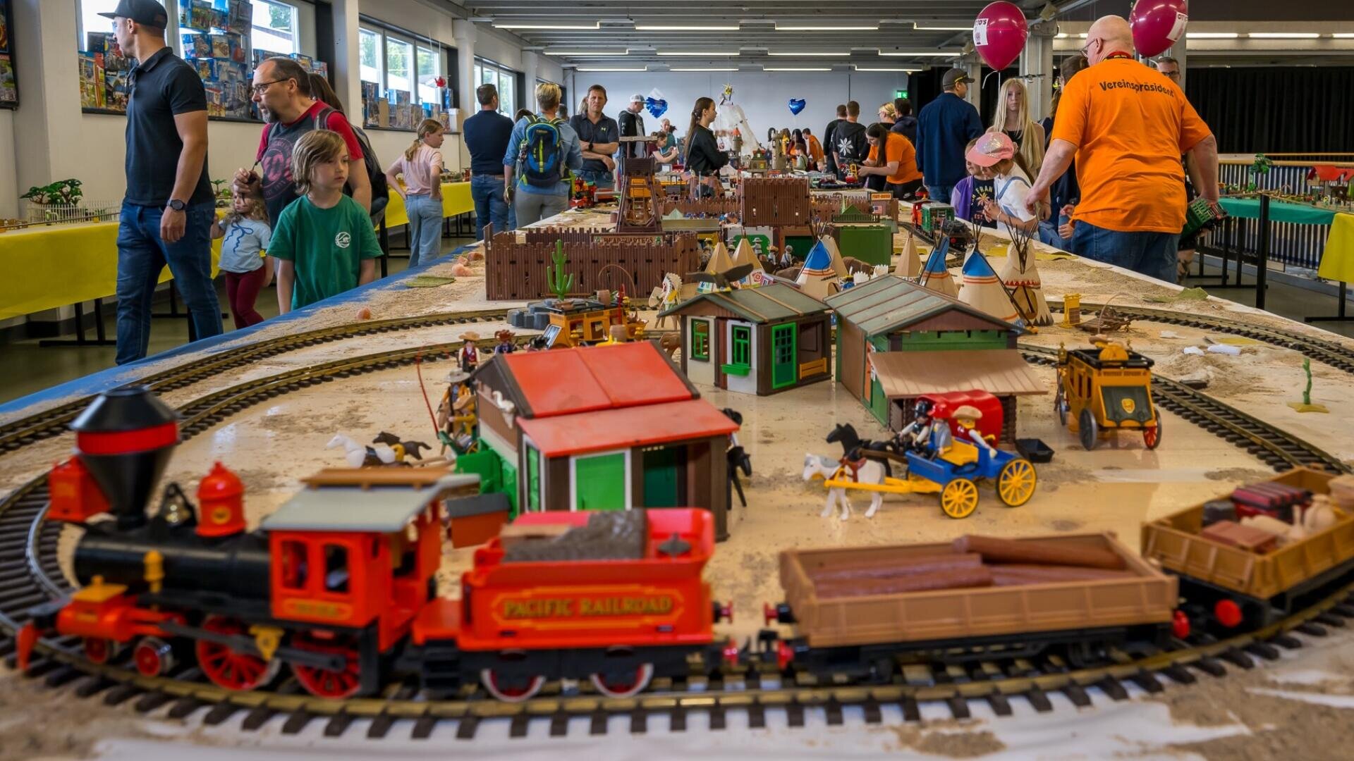 Im Innenbereich ist eine große Tischmodelleisenbahn mit einem roten Spielzeugeisenbahnzug, Schienen, Miniaturgebäuden und Menschen, die herumstehen und die Anlage beobachten, ausgestellt. Kinder und Erwachsene beschäftigen sich mit dem Exponat.