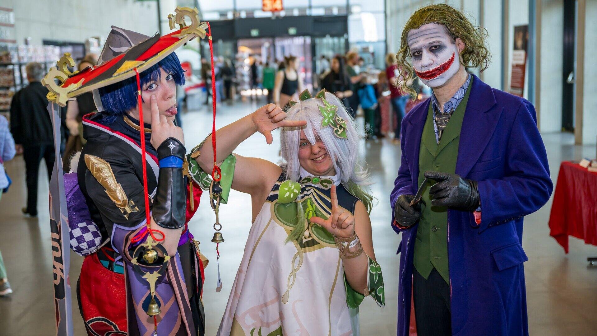 Drei Personen in farbenfrohen und detaillierten Cosplay-Kostümen posieren zusammen in einem Innenraum; eine ist als Joker gekleidet, die beiden anderen als Fantasy-Anime-Figuren, die alle lächeln und spielerische Handgesten machen.