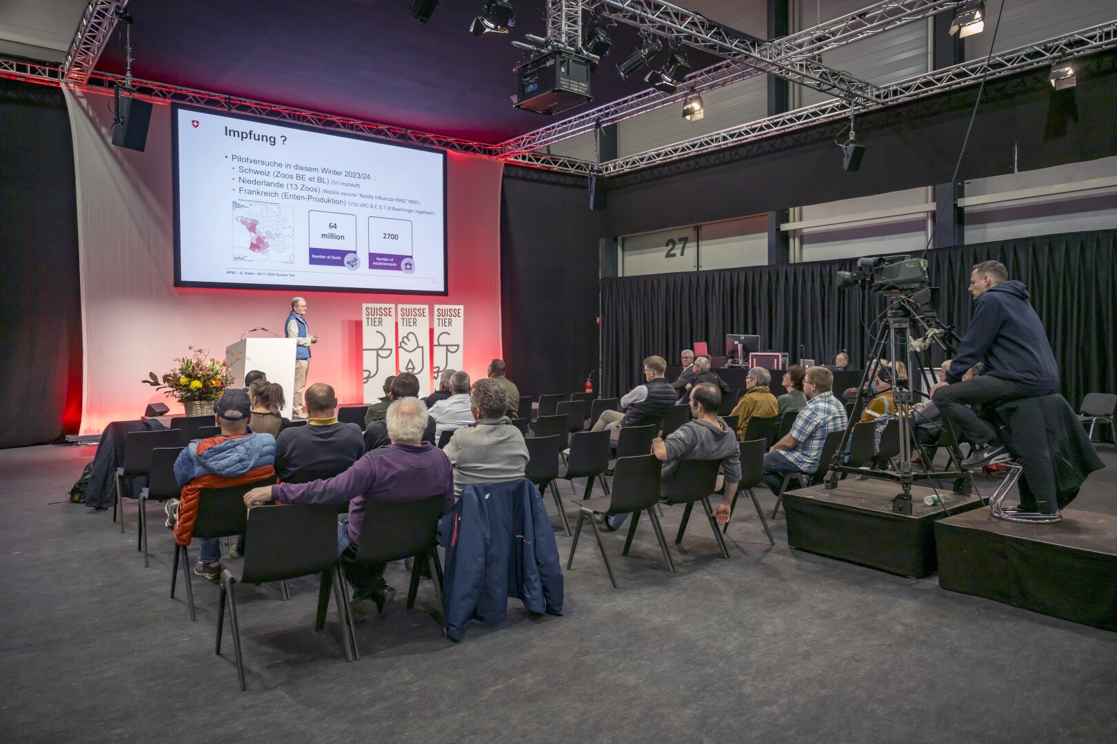 Ein Redner präsentiert einem Publikum in einem Konferenzraum eine Folie zum Thema Impfungen. Ein Kamerateam zeichnet die Veranstaltung auf, und dank der Beleuchtungsanlage über dem Kopf herrscht eine professionelle Atmosphäre.