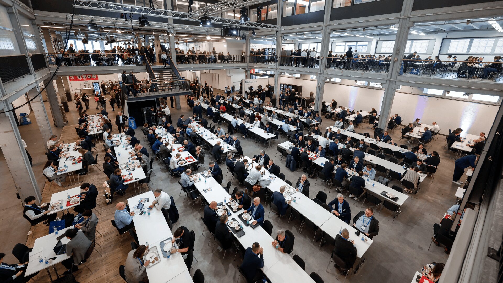 Großer, offener Speisesaal, in dem viele Menschen an langen weißen Tischen sitzen, essen und sich unterhalten; auf einer oberen Ebene sind weitere Sitzplätze sichtbar und der Raum ist hell erleuchtet und weist ein modernes Industriedesign auf.