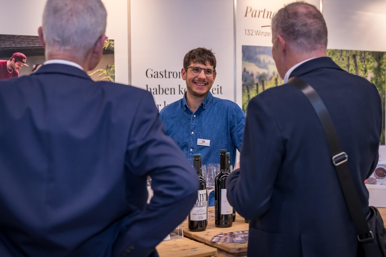Ein lächelnder Mann in einem Jeanshemd steht hinter einem Messestand und unterhält sich mit zwei Männern in Anzügen.