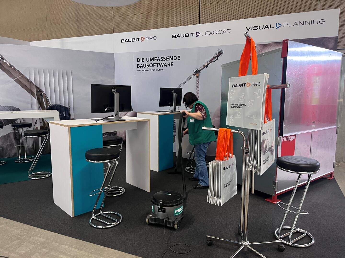Eine Person saugt den Boden in einem modernen Messestand mit Hockern, Computern, Werbetaschen und Schildern für BauBit Pro, Lexocad und Visual Planning Software.