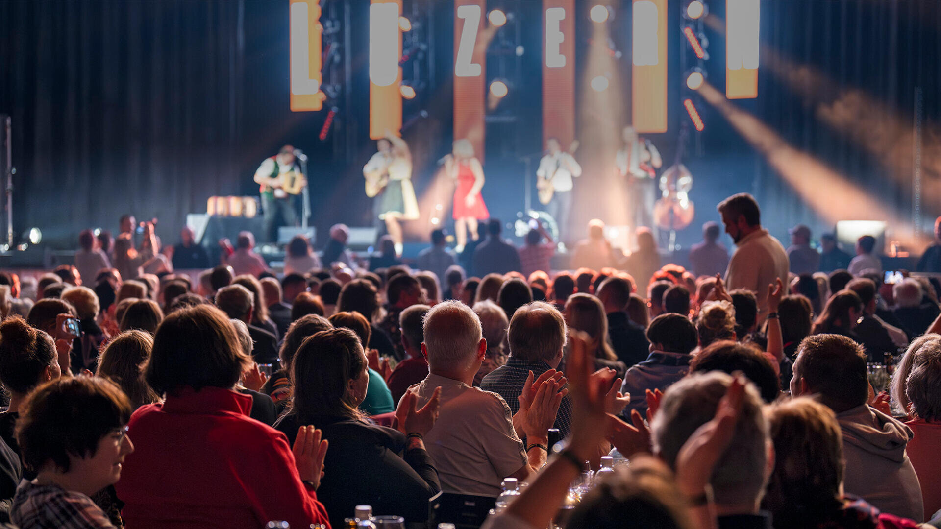 Ein großes Publikum sitzt an Tischen, applaudiert und schaut einer Band zu, die auf einer hell erleuchteten Bühne in einem dunklen Innenraum auftritt. Es herrscht eine lebhafte Atmosphäre, und die Menschen genießen die Konzertatmosphäre.