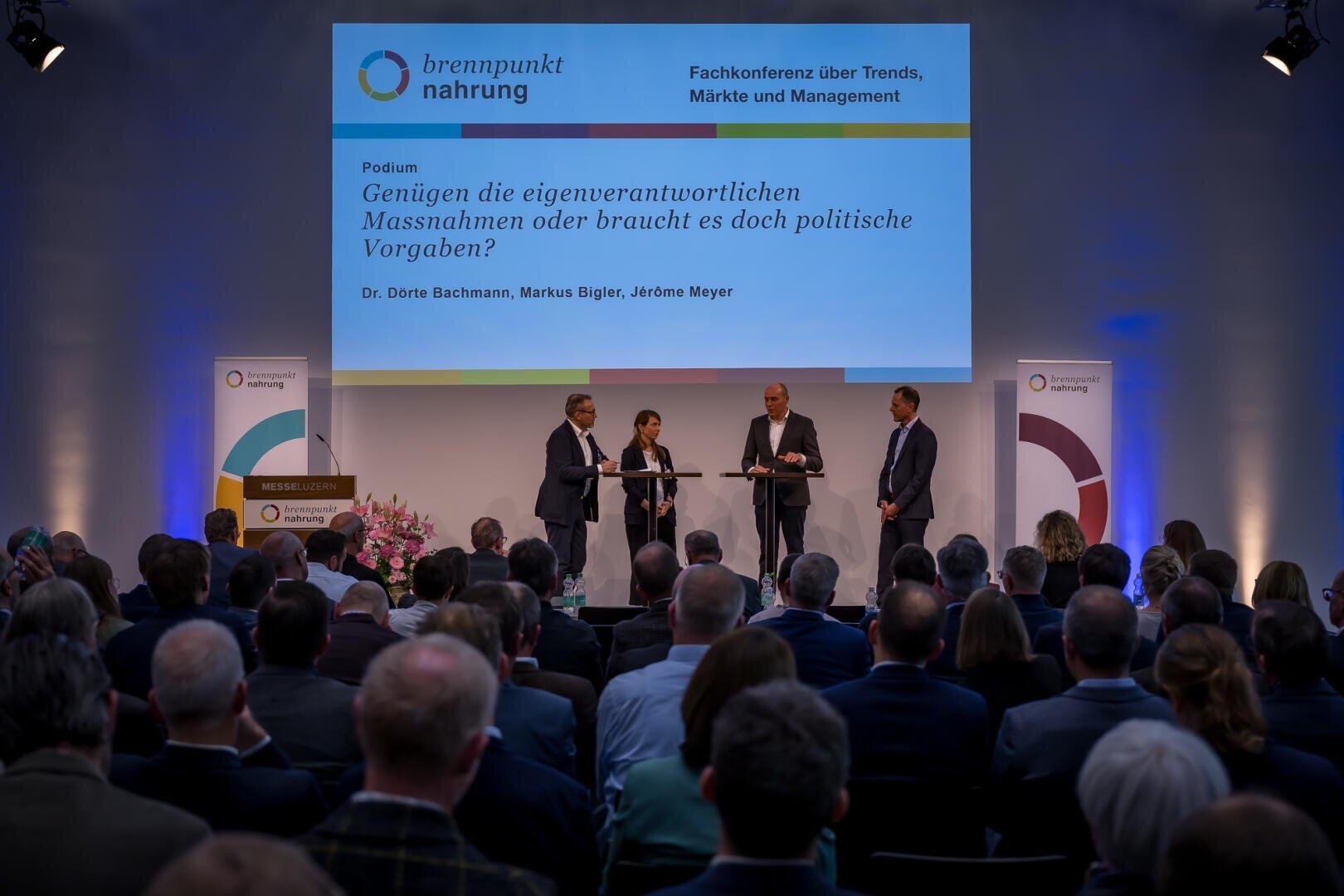 Ein vierköpfiges Gremium steht auf der Bühne und diskutiert unter einem großen Bildschirm, auf dem der Titel der Konferenz angezeigt wird, während das Publikum in einem schwach beleuchteten Hörsaal zuhört. Das Thema der Veranstaltung lautet "Verantwortliches Handeln versus politische Regulierung".