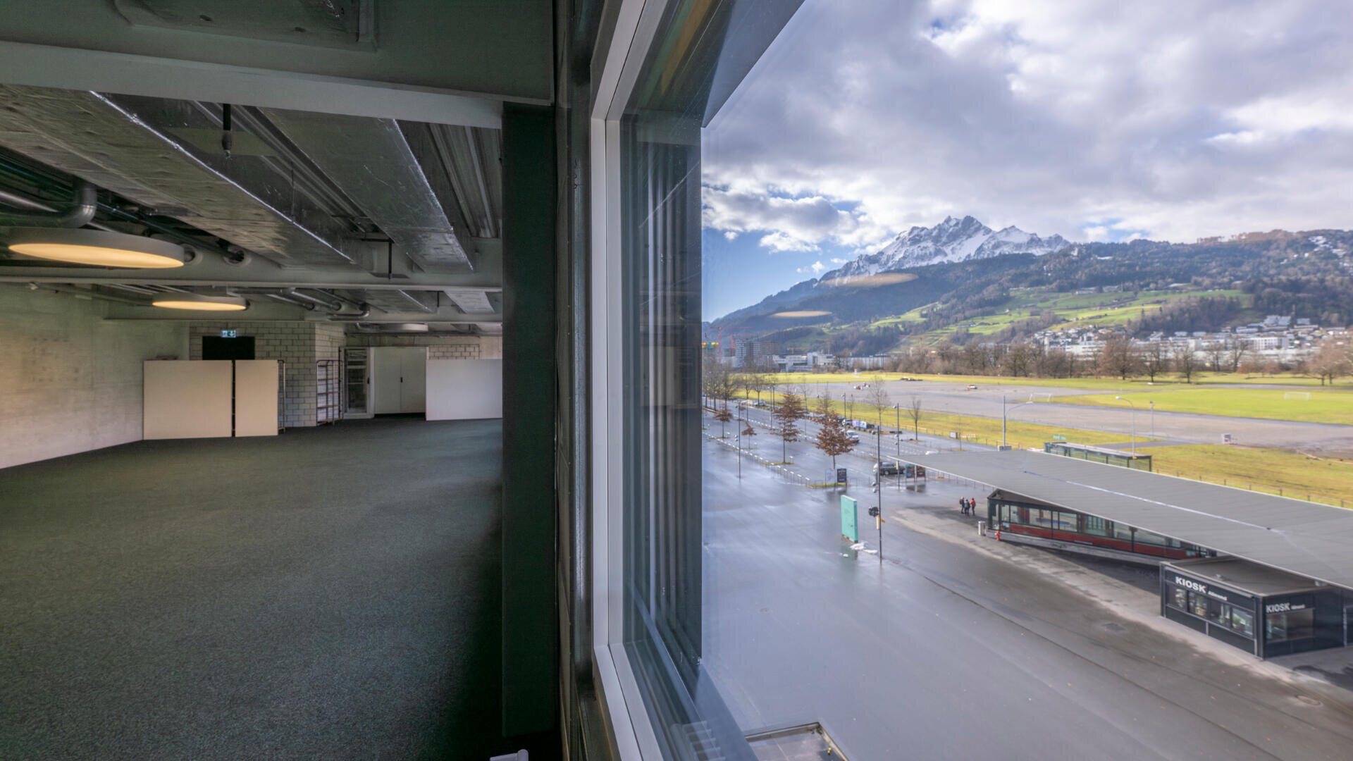 Der Blick durch ein großes Fenster zeigt links einen leeren, modernen Innenraum und rechts eine malerische Außenlandschaft mit Bergen, grünen Feldern und bewölktem Himmel.