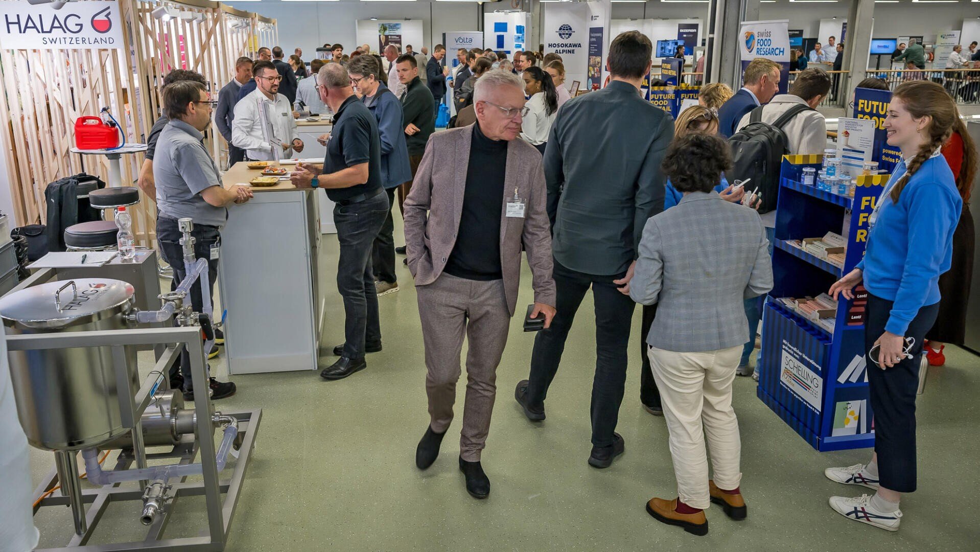 Menschen, die sich in einem hellen Konferenzsaal mit verschiedenen Unternehmensständen, Geräten und Informationsdisplays austauschen.