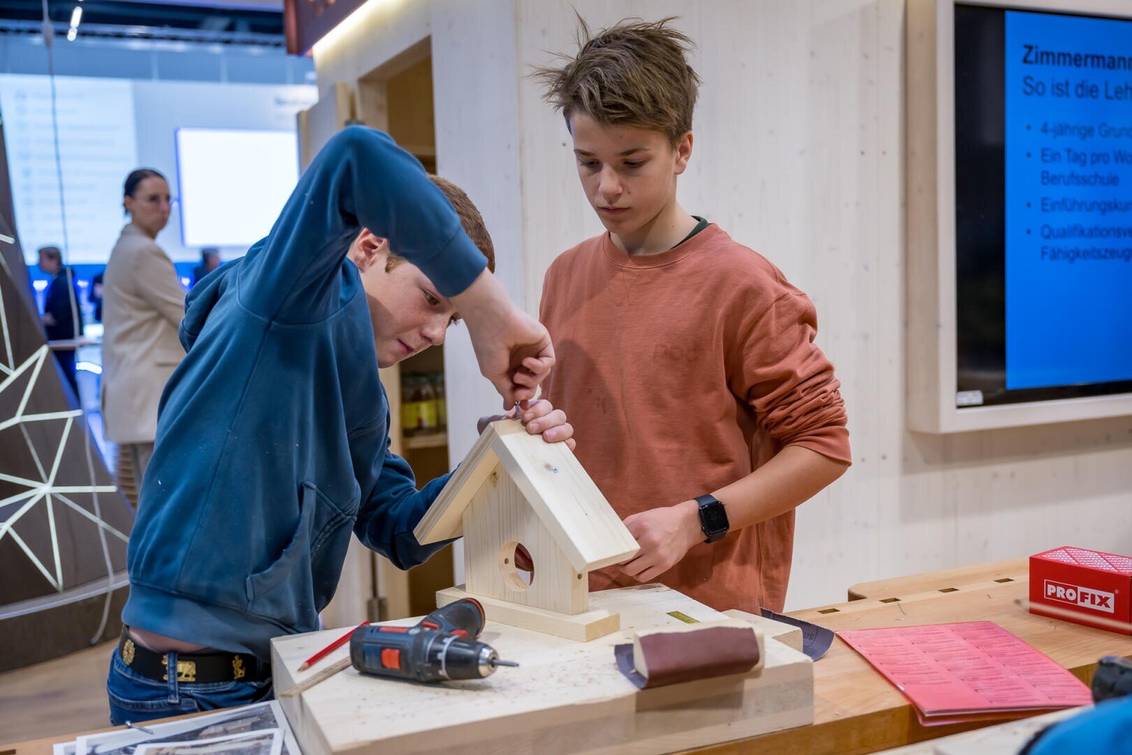 Zwei Jungen bauen gemeinsam an einem Tisch ein Vogelhaus aus Holz zusammen. Einer hält das Dach, während der andere einen Schraubenzieher benutzt. Werkzeuge und Materialien liegen verstreut auf dem Tisch, und im Hintergrund ist ein Informationsdisplay zu sehen.