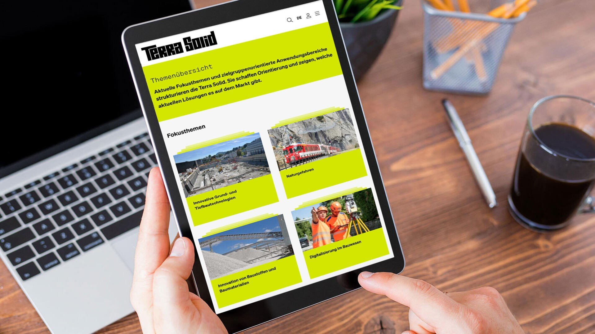 Eine Person hält ein Tablet mit der Terra Sound-Website und hervorgehobenen Nachrichtenabschnitten in der Hand. Daneben liegen ein Kaffeeglas, Bleistifte und Papier auf einem Holzschreibtisch.