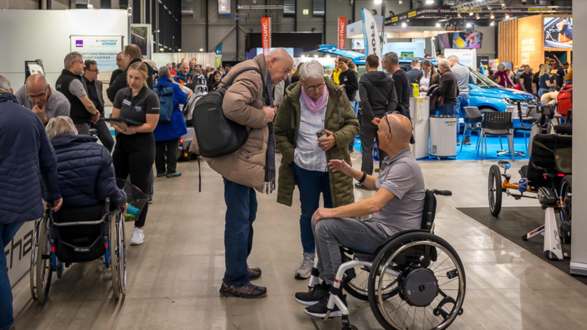 Auf einer geschäftigen Indoor-Expo herrscht reger Austausch zwischen den Menschen. Mehrere Rollstuhlfahrer unterhalten sich zwischen Ständen, an denen Produkte zur Barrierefreiheit und adaptive Ausrüstung ausgestellt sind, mit den Ausstellern und untereinander.