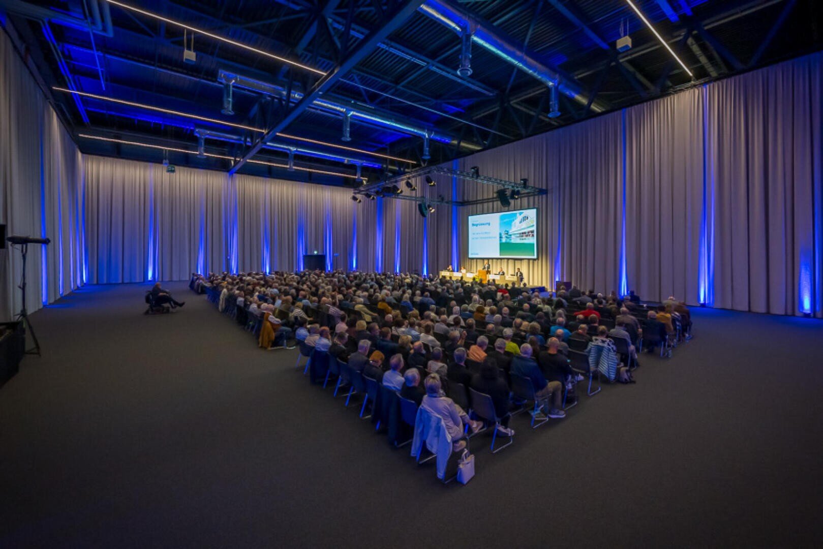 In einem geräumigen, modernen Konferenzsaal mit blauer Beleuchtung und hohen Decken sitzt ein großes Publikum in Reihen vor einer Bühne mit Rednern und einer Projektionsfläche.