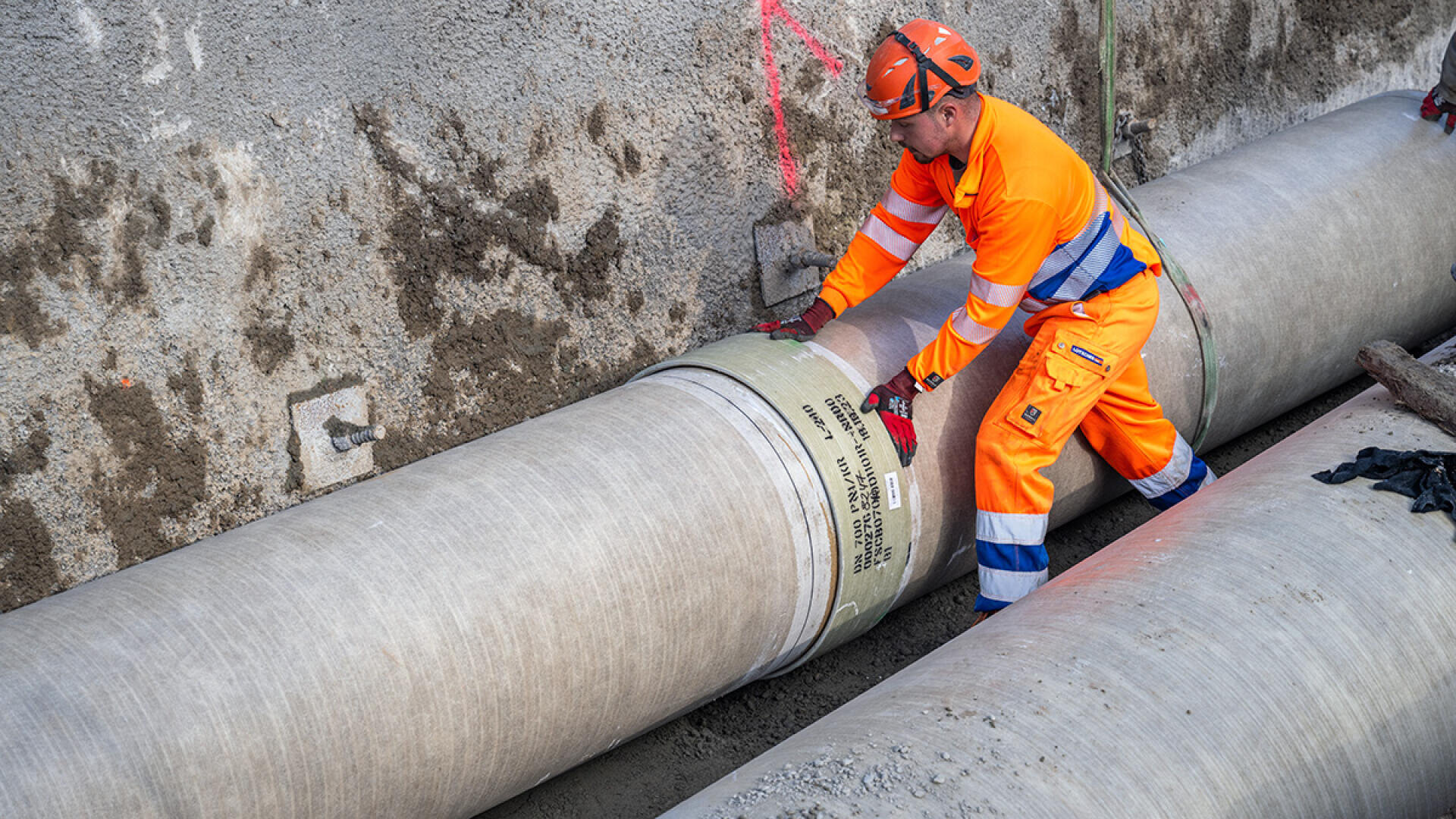 Ein Bauarbeiter in orangefarbener Schutzkleidung sichert ein großes Betonrohr in einem Graben auf einer Baustelle, umgeben von Erde und Betonwänden.