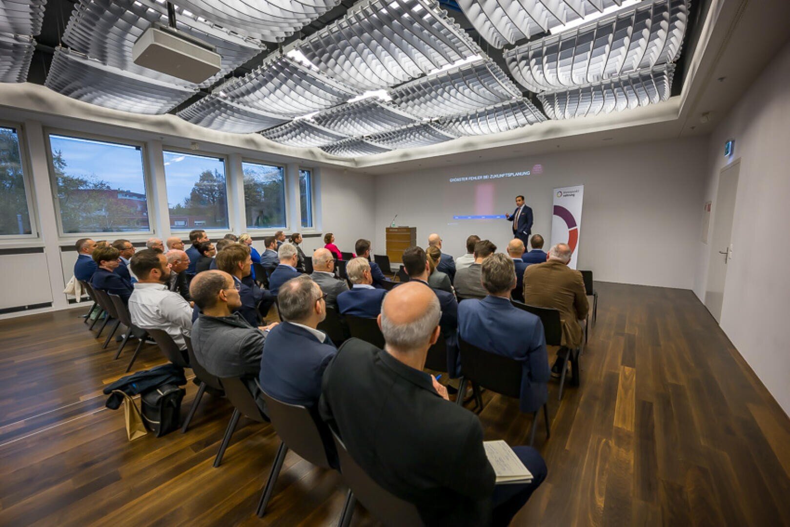 Eine Gruppe von Personen in Geschäftskleidung sitzt in einem modernen Konferenzraum und hört einem Redner zu, der im vorderen Teil des Raums Folien präsentiert. Große Fenster lassen natürliches Licht herein, und die Decke ist mit einem einzigartigen Muster versehen.