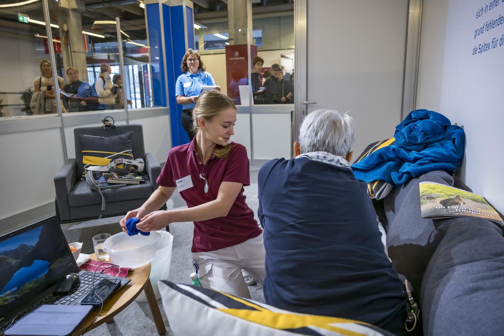 Eine Mitarbeiterin des Gesundheitswesens in einem kastanienbraunen Polohemd wäscht die Hände einer älteren Person, die auf einem Sofa sitzt.