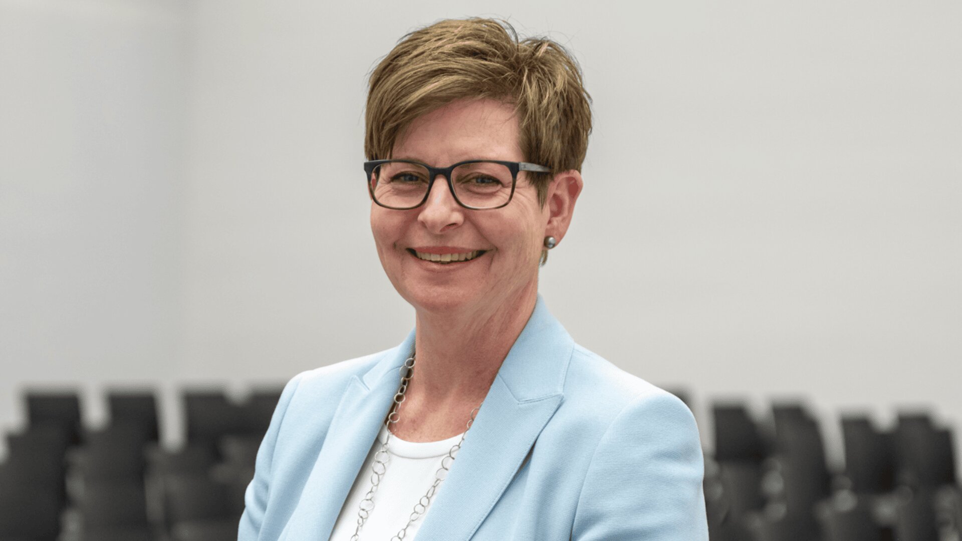 Eine Frau mit kurzen braunen Haaren und Brille lächelt, sie trägt einen hellblauen Blazer über einem weißen Oberteil. Sie steht vor einer Reihe leerer schwarzer Stühle in einem hellen Raum.