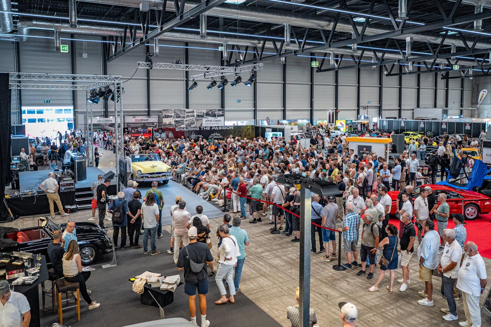 Eine große Menschenmenge auf einer Autoausstellung in einer Halle betrachtet einen ausgestellten gelben Oldtimer. Die Besucher stehen und sitzen, und in der Halle sind verschiedene Oldtimer und Ausstellungsstücke zu sehen.
