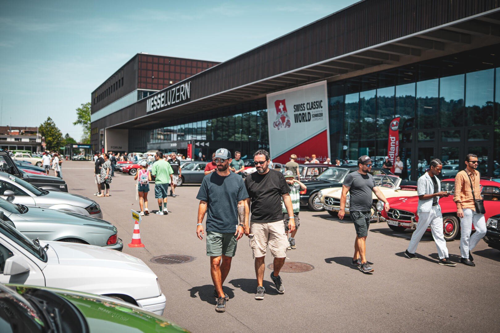 An einem sonnigen Tag schlendern die Besucher während der „Swiss Classic World“ zwischen den vor der Messe Luzern geparkten Oldtimern umher. Es herrscht reges Treiben: Die Menschenmengen bewundern die Fahrzeuge, und an dem Gebäude sind Banner zu sehen.