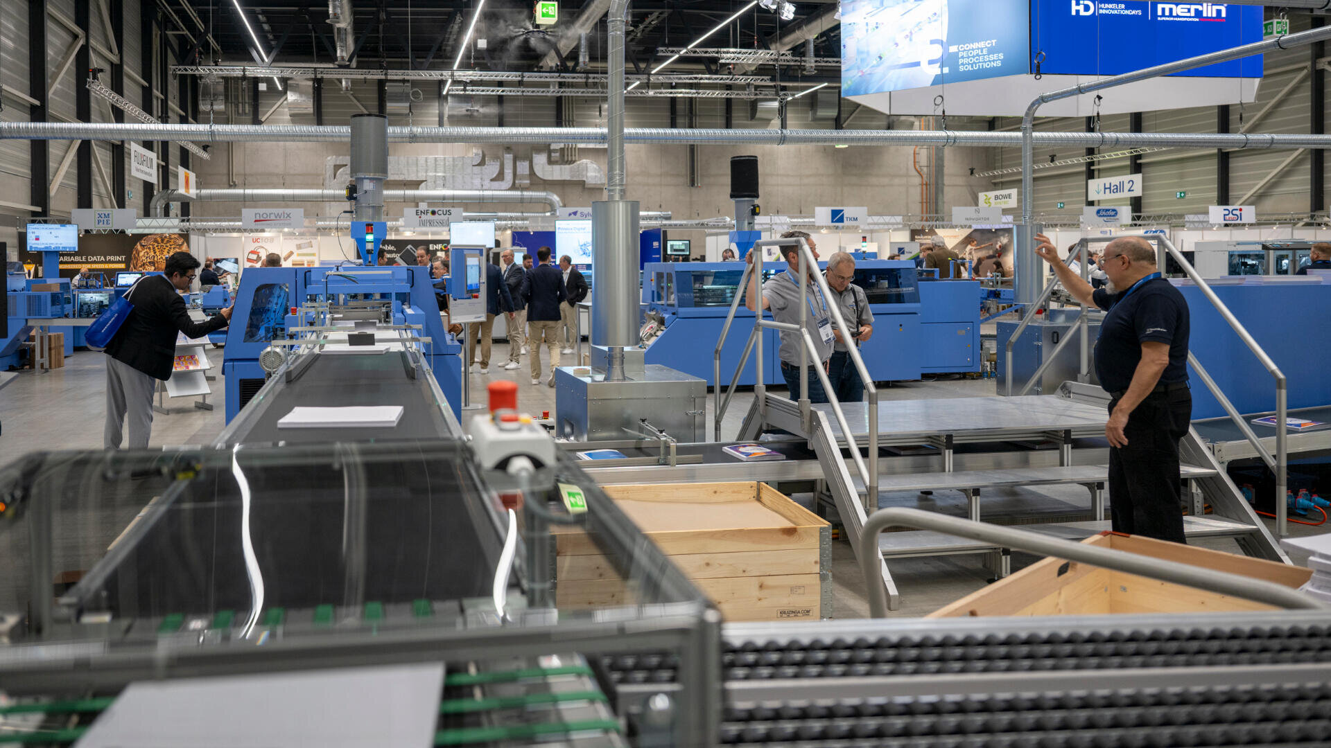 Auf einer Messe arbeiten Menschen in einer geräumigen, hellen Messehalle an großen blauen Industriemaschinen. Einige interagieren mit den Geräten, während andere beobachten oder sich in kleinen Gruppen unterhalten.