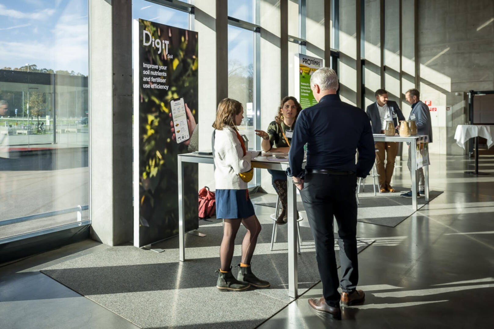 In einem modernen, sonnendurchfluteten Konferenzraum mit großen Glasfenstern wird an hohen Tischen diskutiert und vernetzt. Mehrere Poster und Banner zum Thema digitale Innovation sind in dem Raum verteilt.